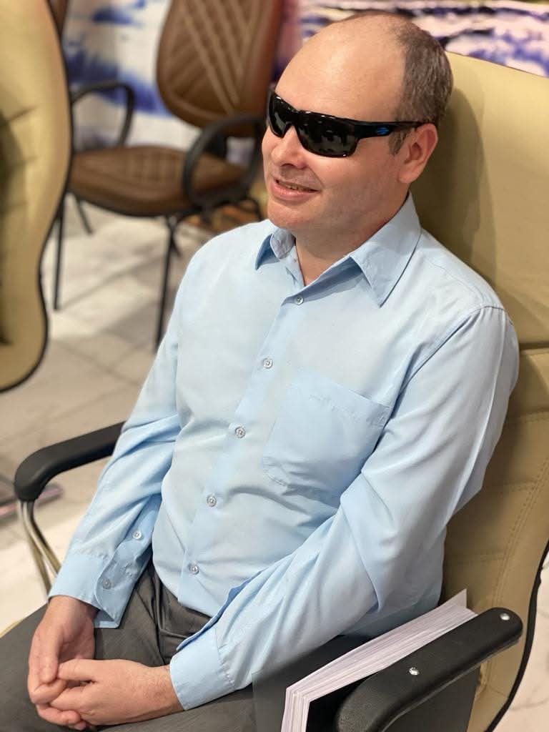 Homem adulto de pele clara sentado em uma poltrona bege. Ele usa óculos escuros grandes com armação preta e está sorrindo levemente, com a cabeça um pouco voltada para a direita. Veste camisa social azul-clara, bem passada, abotoada até o colarinho, e calça social cinza.